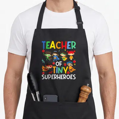 Superheroe tiny chef apron - Teacher of Tiny Superheroes text with six costumed kids, apples, and lightning bolts — shown on a black cotton canvas apron against a white background