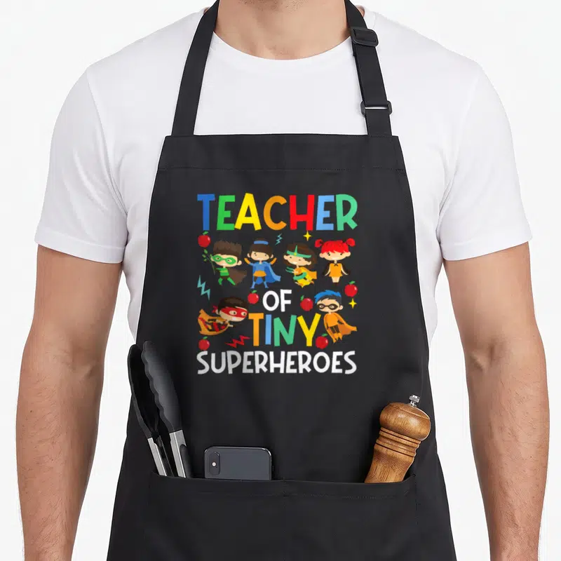 Superheroe tiny chef apron - Teacher of Tiny Superheroes text with six costumed kids, apples, and lightning bolts — shown on a black cotton canvas apron against a white background