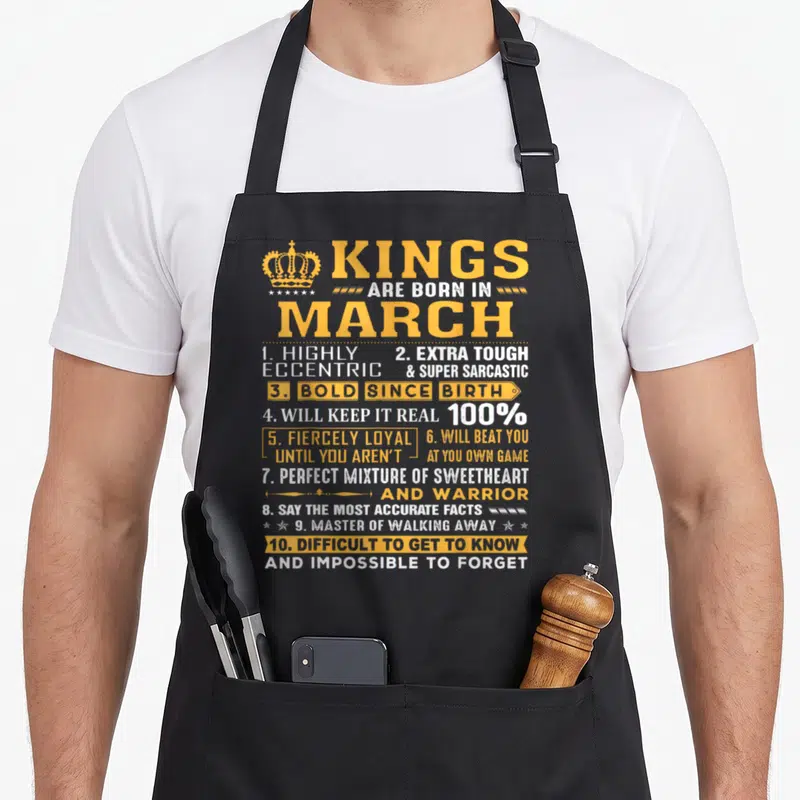 March king apron - Orange and white typography listing ten traits of Kings Born In March with a crown icon — shown on a black cotton canvas apron against a white background