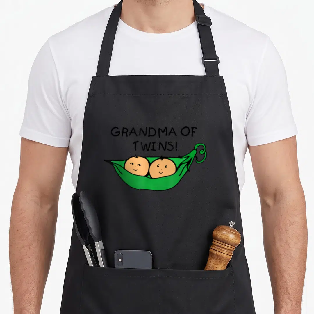Peas grandma apron - Text GRANDMA OF TWINS above cartoon green pea pod with two babies — shown on a black cotton canvas apron against a white background