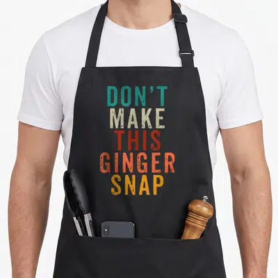 Ginger snap aprons - Stacked text reading DON'T MAKE THIS GINGER SNAP in teal beige red orange and yellow — shown on a black cotton canvas apron against a white background