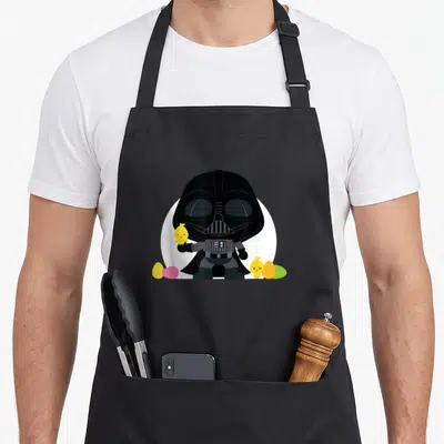 Kawaii egg apron - Cartoon Darth Vader holding a yellow chick with four colorful eggs at his feet — shown on a black cotton canvas apron against a white background