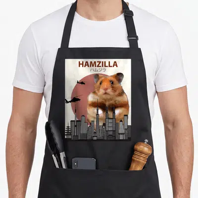 Hamzilla crazy hamster man apron - Giant brown hamster behind city skyline with red sun and two helicopters flying left — shown on a black cotton canvas apron against a white background
