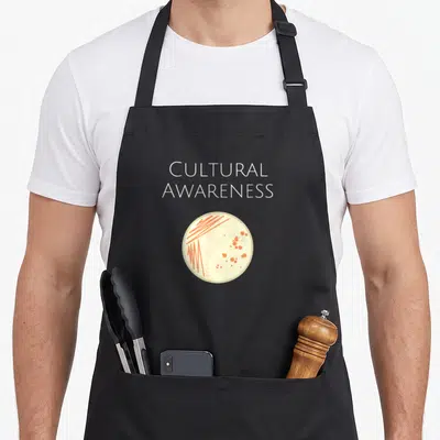 Microbiology microbiologist apron - CULTURAL AWARENESS text above petri dish with orange bacterial streaks — shown on a black cotton canvas apron against a white background
