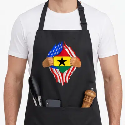 Ghanaian blood apron - Two brown hands tearing a US flag to reveal the Ghana flag with red yellow green blue and white — shown on a black cotton canvas apron against a white background