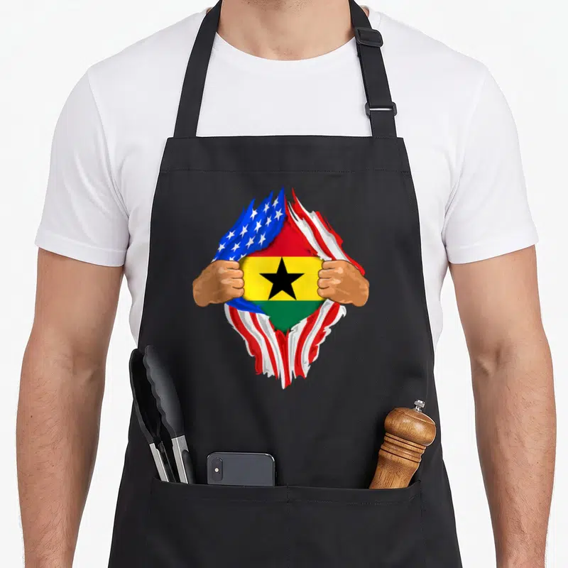 Ghanaian blood apron - Two brown hands tearing a US flag to reveal the Ghana flag with red yellow green blue and white — shown on a black cotton canvas apron against a white background