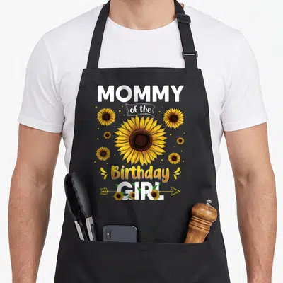 Family sunflower apron - MOMMY of the Birthday GIRL text with large yellow sunflower and scattered dots — shown on a black cotton canvas apron against a white background