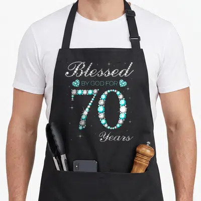Blessed god 70th birthday apron - Rhinestone text Blessed BY GOD FOR 70 Years in silver and cyan with heart accents — shown on a black cotton canvas apron against a white background