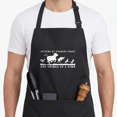 Animal driving aprons - White line art of jumping cow, goat, rooster, dog, and cat under humorous text on black fabric — shown on a black cotton canvas apron against a white background