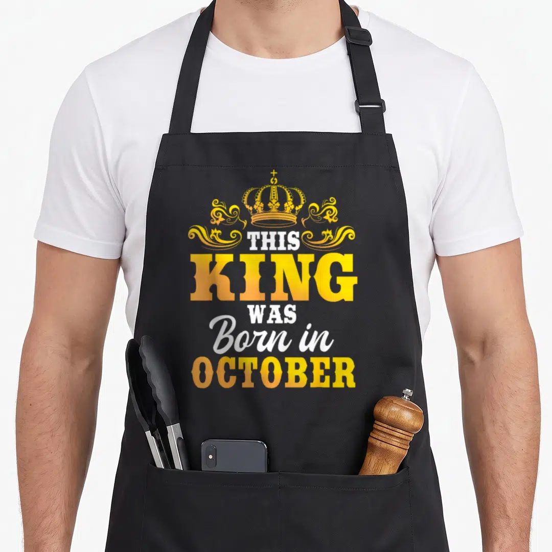 Celebration king apron - Crown and THIS KING WAS Born in OCTOBER text in yellow orange white — shown on a black cotton canvas apron against a white background