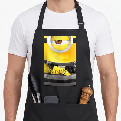 Despicable tattoo apron - Realistic minion with crossed arms and GRU LIFE tattoo text on yellow arm — shown on a black cotton canvas apron against a white background