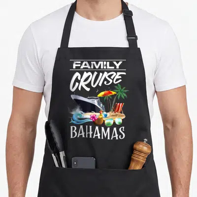 Bahamas family aprons - FAMILY CRUISE BAHAMAS text with cruise ship, beach umbrella, coconut drink, and sunglasses — shown on a black cotton canvas apron against a white background