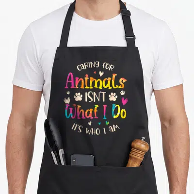 Caring animal apron - Colorful text caring for Animals ISN'T What I Do ITS WHO I AM with paw prints and hearts — shown on a black cotton canvas apron against a white background