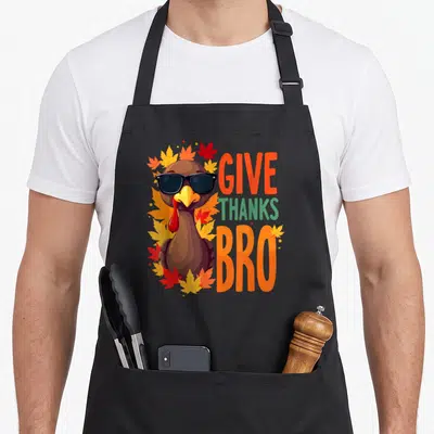 Give thankful apron - Cool turkey with sunglasses and autumn leaves beside stacked GIVE THANKS BRO text — shown on a black cotton canvas apron against a white background