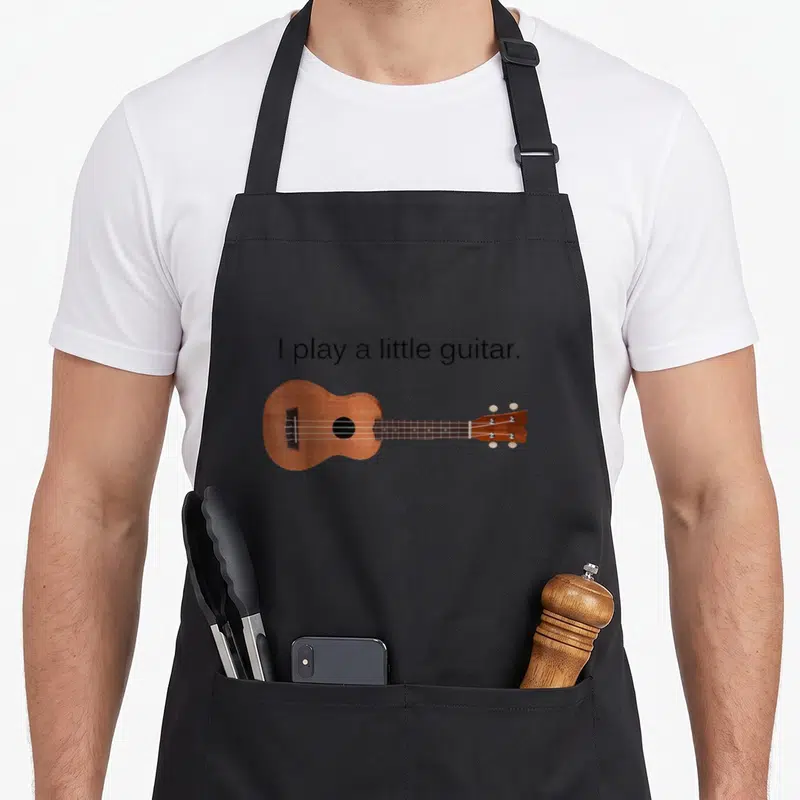 Guitar ukulele apron - Brown ukulele with four strings below black text reading I play a little guitar — shown on a black cotton canvas apron against a white background