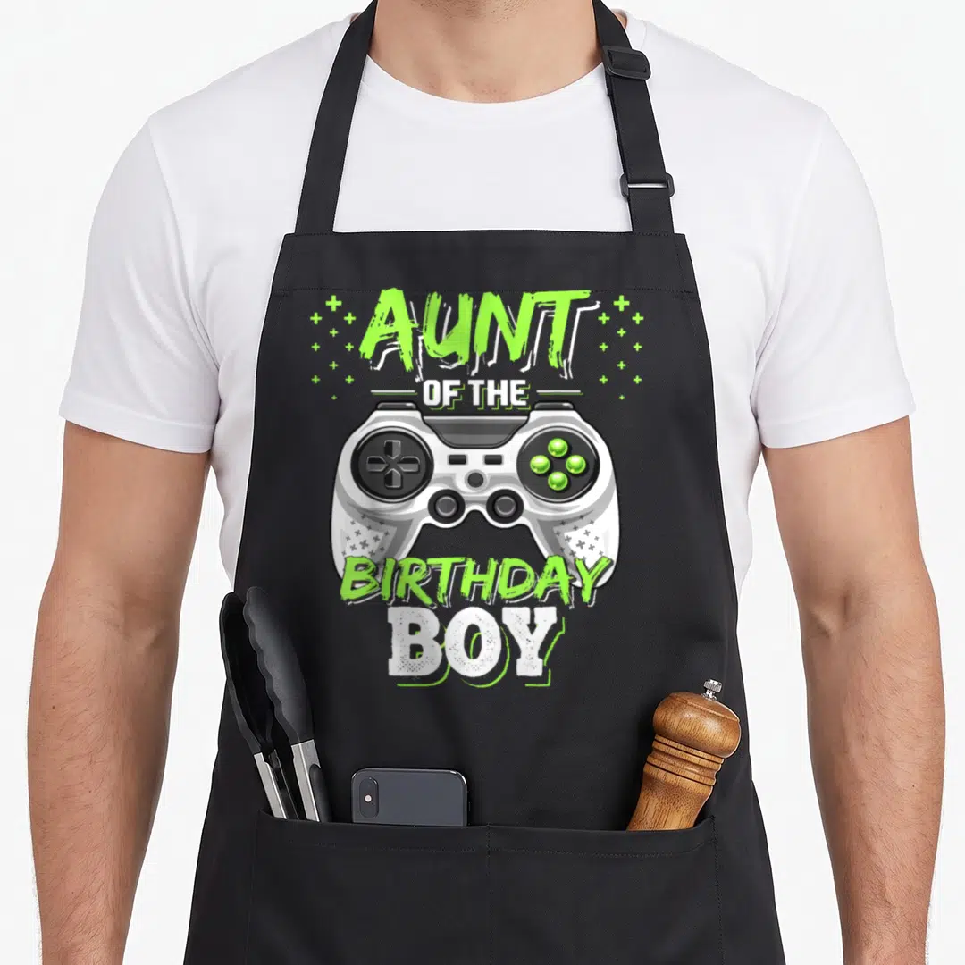 Aunt video game apron - AUNT OF THE BIRTHDAY BOY text with white game controller and lime green plus signs — shown on a black cotton canvas apron against a white background