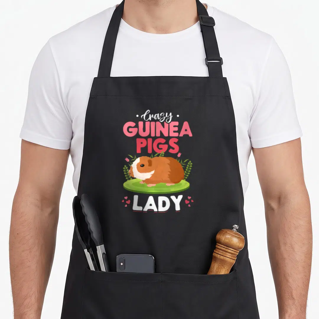 Cute guinea animal apron - Crazy Guinea Pigs Lady text with brown guinea pig on green grass and pink hearts — shown on a black cotton canvas apron against a white background