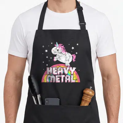 Ironic heavy metal apron - Cartoon unicorn on rainbow with HEAVY METAL text and white stars — shown on a black cotton canvas apron against a white background