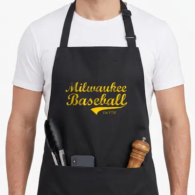 Milwaukee wisconsin apron - Yellow distressed script text reading Milwaukee Baseball Est 1970 with a decorative swoosh — shown on a black cotton canvas apron against a white background