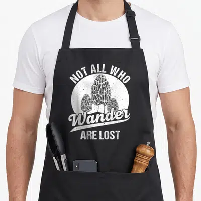 Hunter foraging apron - Three white morel mushrooms in a circle with text Not All Who Wander Are Lost — shown on a black cotton canvas apron against a white background