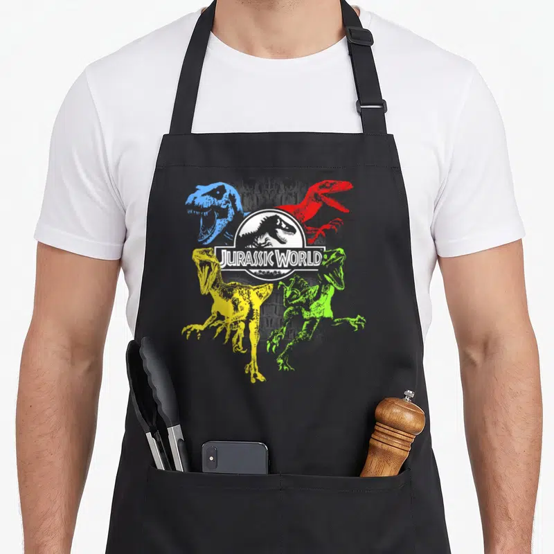 Primary t-rex apron - JURASSIC WORLD logo with blue, red, yellow, green cartoon dinosaurs — shown on a black cotton canvas apron against a white background