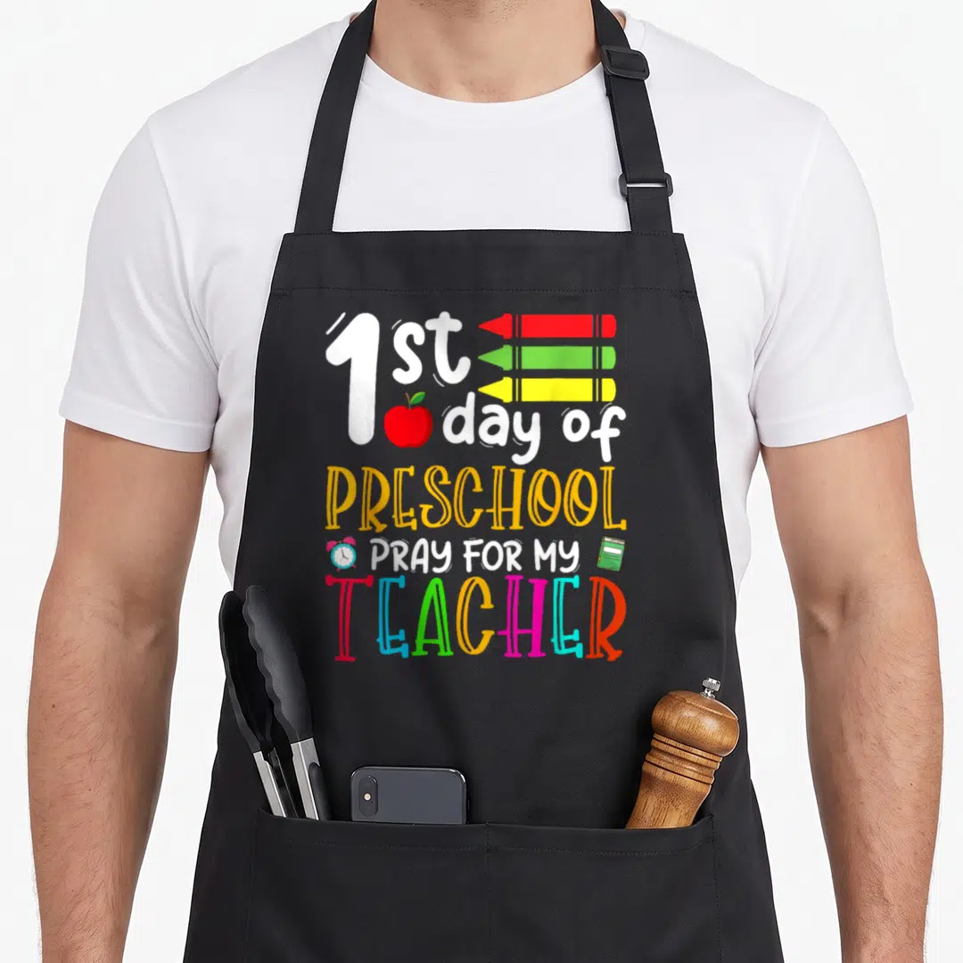 Pray preschool apron - Stacked text 1st day preschool pray teacher with three crayons red apple and alarm clock — shown on a black cotton canvas apron against a white background