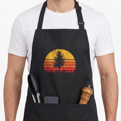 Sun minimalist apron - Grey pine tree silhouette over horizontal yellow orange and red stripes — shown on a black cotton canvas apron against a white background