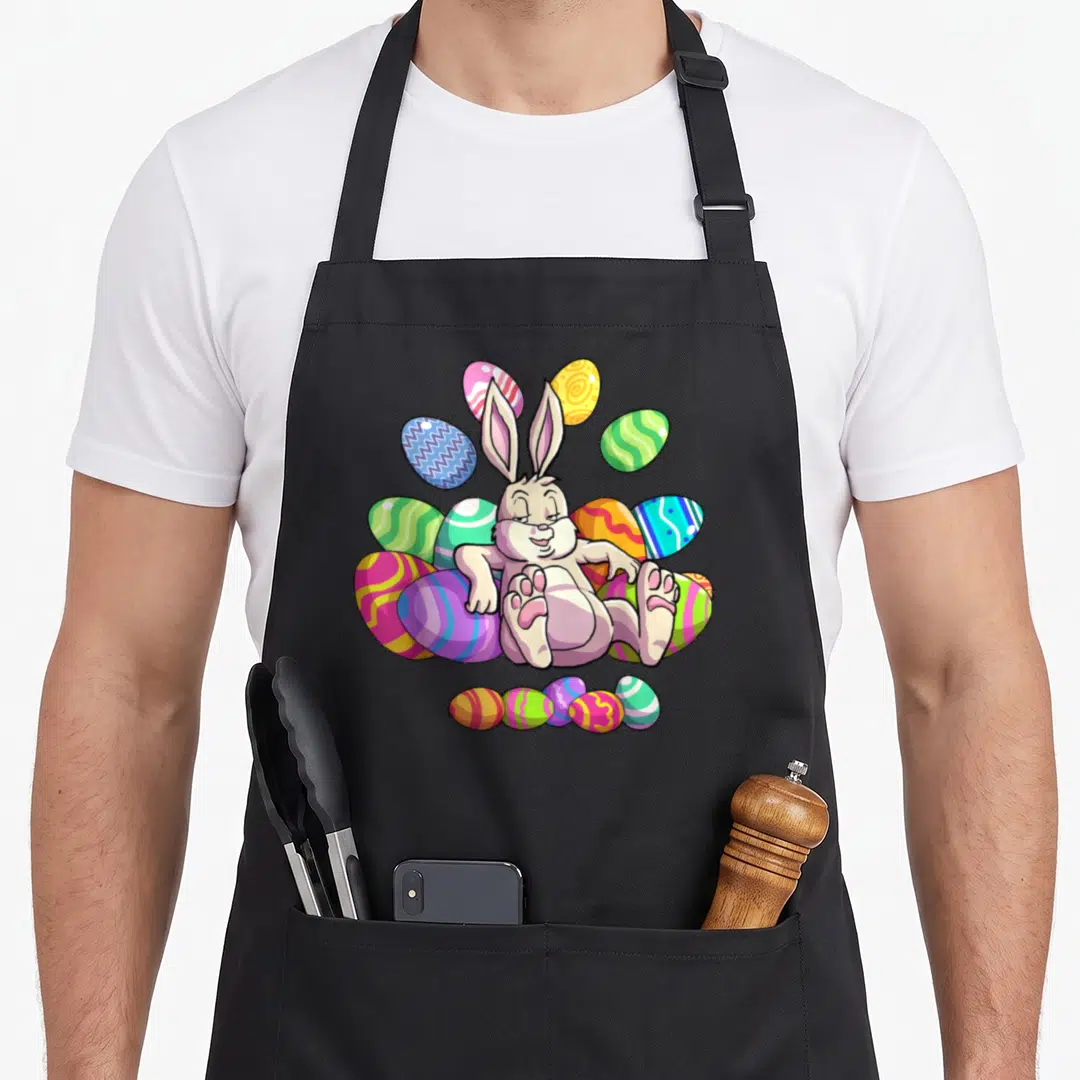 Funny easter bunny apron - Cartoon bunny sitting among purple pink yellow green and blue eggs — shown on a black cotton canvas apron against a white background