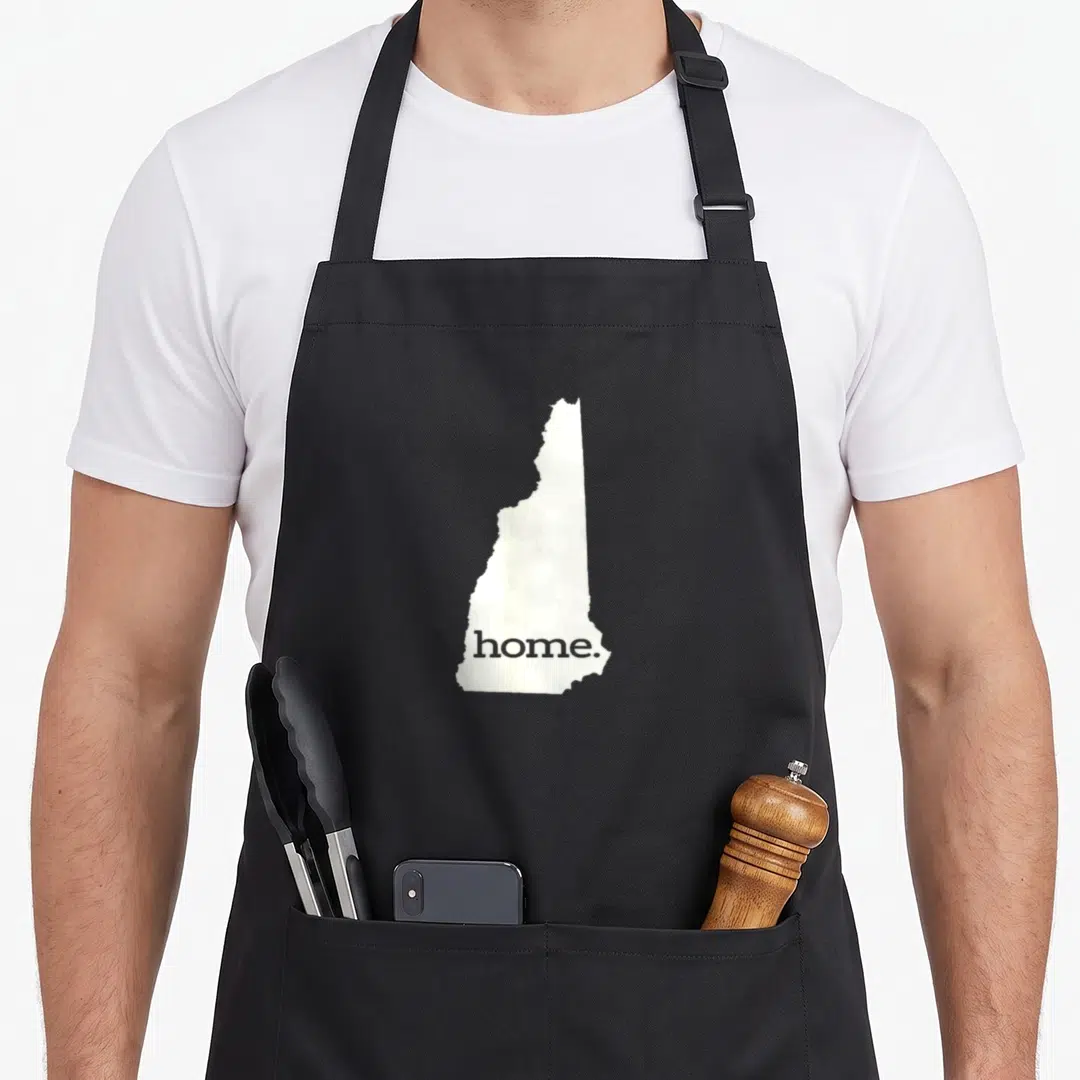 Funny hampshire boy apron - White New Hampshire state silhouette containing the grey word home. inside the bottom — shown on a black cotton canvas apron against a white background