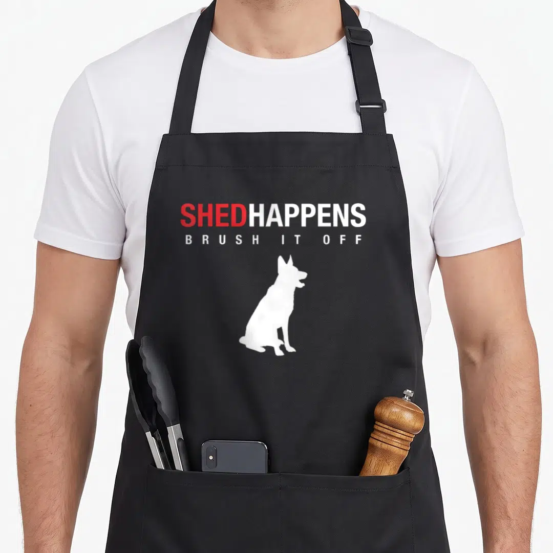Shed dog mom apron - Red and white text reading Shed Happens Brush It Off above a sitting white dog silhouette — shown on a black cotton canvas apron against a white background