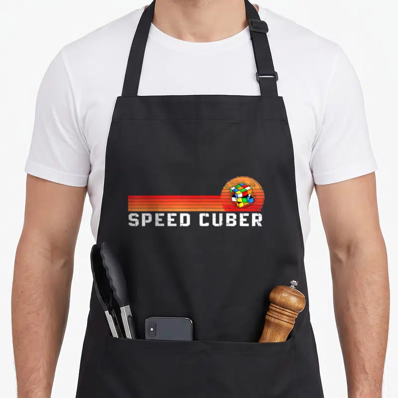 Math speed girl apron - Retro red and orange sun behind a Rubik's cube with SPEED CUBER text below — shown on a black cotton canvas apron against a white background