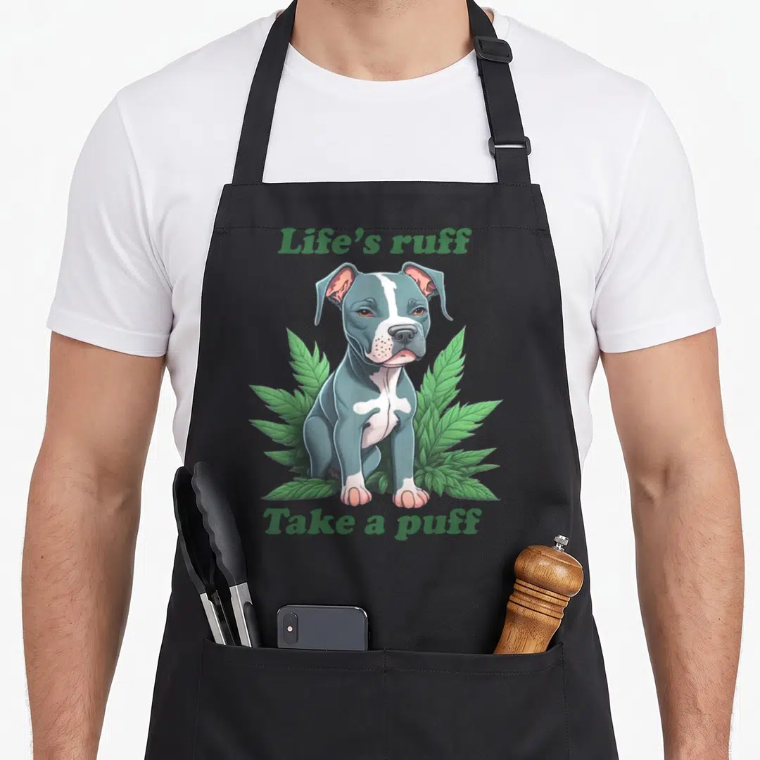 Ruff puff apron - Seated blue-grey dog surrounded by green leaves with Life's ruff Take a puff text — shown on a black cotton canvas apron against a white background
