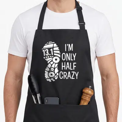 Funny runner marathon apron - White shoe print with 13.1 text and I'M ONLY HALF CRAZY stacked lettering — shown on a black cotton canvas apron against a white background