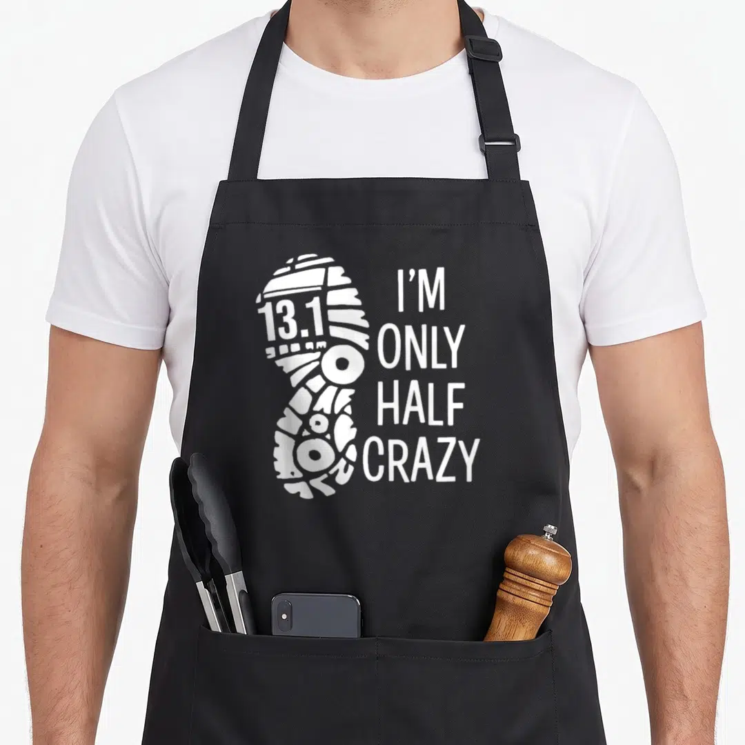 Funny runner marathon apron - White shoe print with 13.1 text and I'M ONLY HALF CRAZY stacked lettering — shown on a black cotton canvas apron against a white background