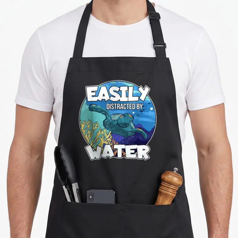 Easily snorkeling quotes apron - Cartoon sea turtle and yellow coral in blue circle with white text easily distracted — shown on a black cotton canvas apron against a white background