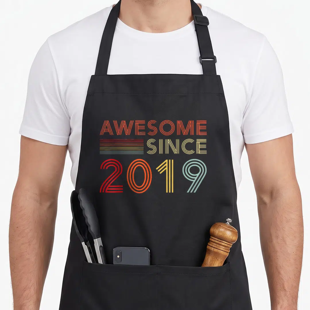 Bday decoration dad and son aprons - Retro striped text reading AWESOME SINCE 2019 in orange red beige yellow and teal — shown on a black cotton canvas apron against a white background