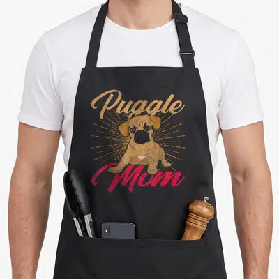 Pet mother's day aprons - Distressed tan Puggle illustration with red Mom text on sunburst background — shown on a black cotton canvas apron against a white background