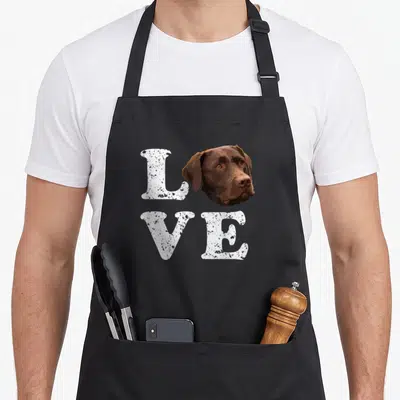 Chocolate labrador apron - Brown dog head replacing O in distressed white L VE text — shown on a black cotton canvas apron against a white background