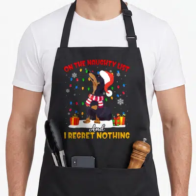 Santa dachshund apron - Cartoon dachshund in Santa hat surrounded by lights and gifts with naughty list text — shown on a black cotton canvas apron against a white background