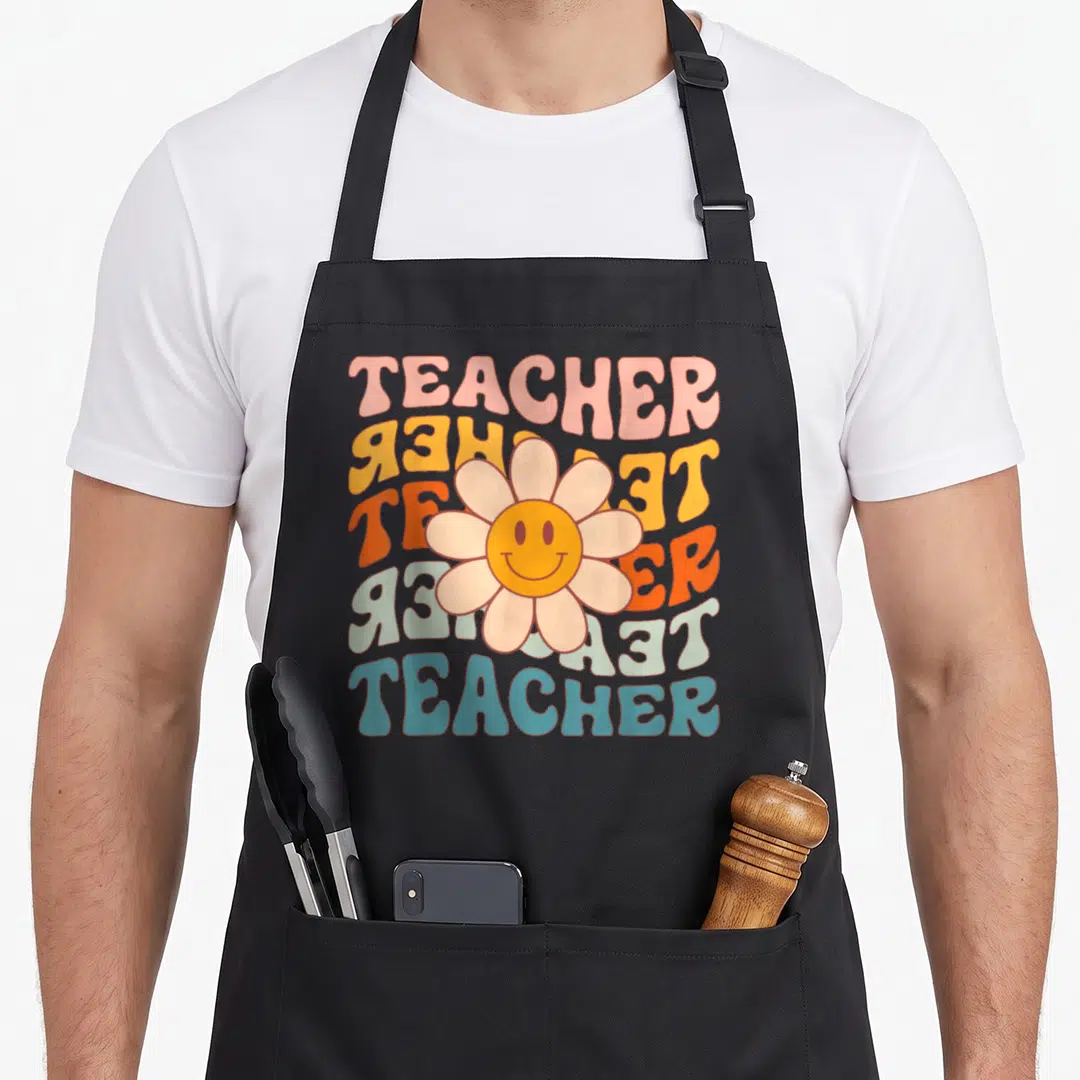 Teacher daisy apron - Retro smiley flower over six wavy TEACHER text lines in pink, yellow, orange, blue, and peach — shown on a black cotton canvas apron against a white background