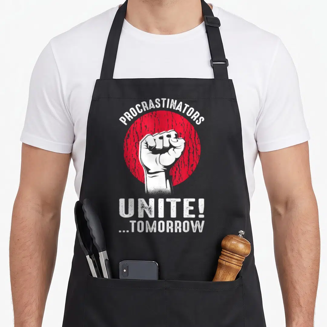 Sarcasm procrastinator apron - White fist inside red circle with text Procrastinators Unite Tomorrow in distressed font — shown on a black cotton canvas apron against a white background