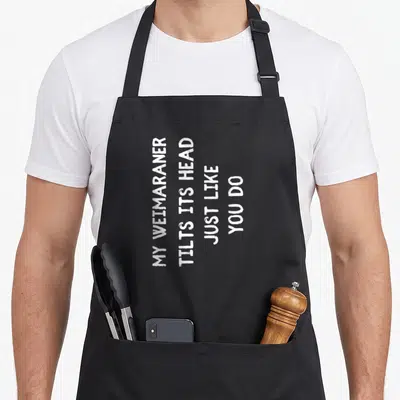 Funny weimaraner dog apron - White vertical text reading My Weimaraner Tilts Its Head Just Like You Do — shown on a black cotton canvas apron against a white background