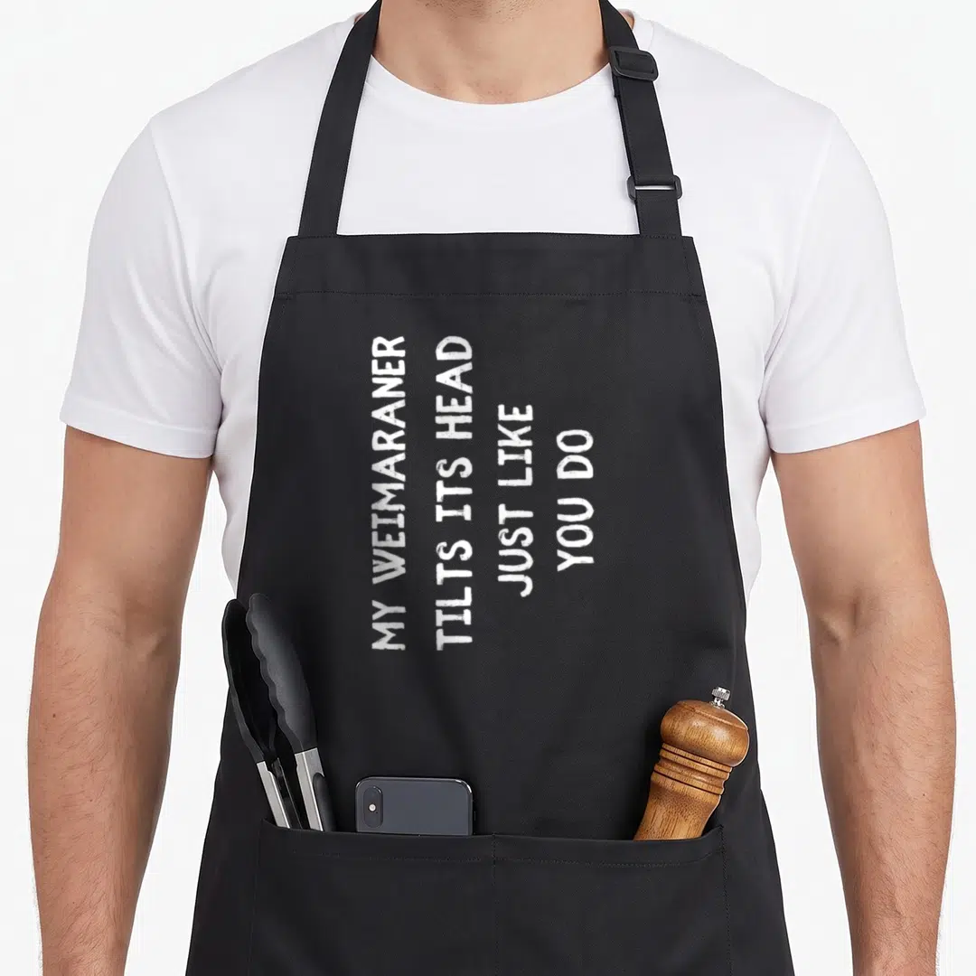 Funny weimaraner dog apron - White vertical text reading My Weimaraner Tilts Its Head Just Like You Do — shown on a black cotton canvas apron against a white background
