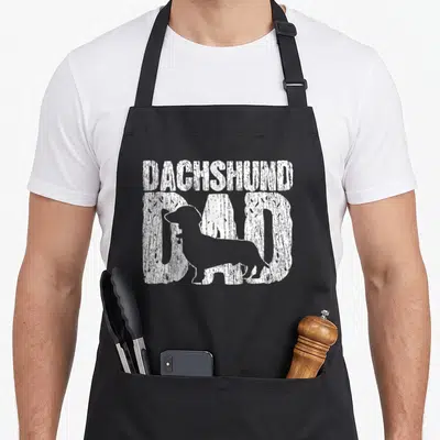 Father dachshund apron - White distressed DAD text with dachshund silhouette overlapping letters — shown on a black cotton canvas apron against a white background