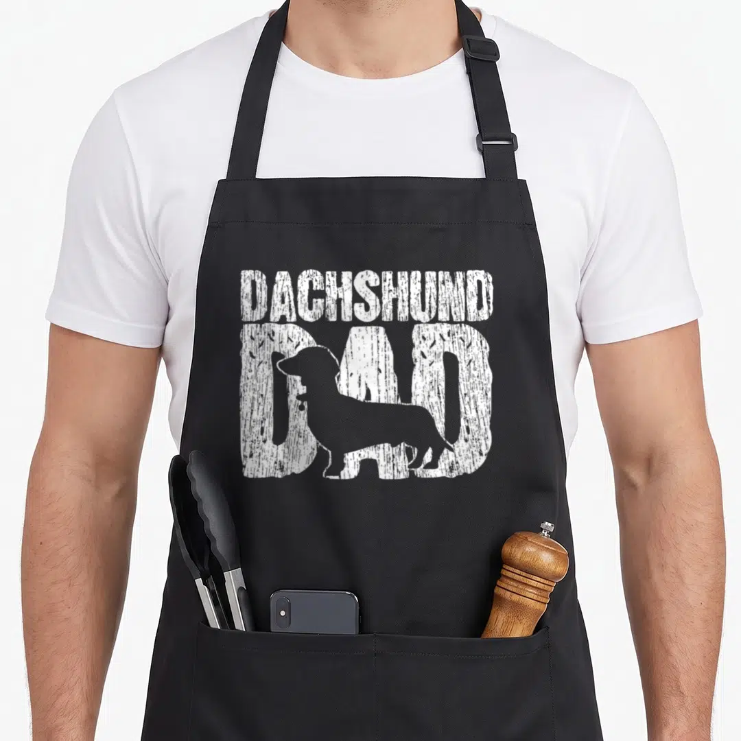 Father dachshund apron - White distressed DAD text with dachshund silhouette overlapping letters — shown on a black cotton canvas apron against a white background