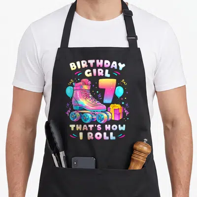 Number birthday apron - Pink roller skate, rainbow number 7, blue balloons, purple gift box, birthday pun text — shown on a black cotton canvas apron against a white background