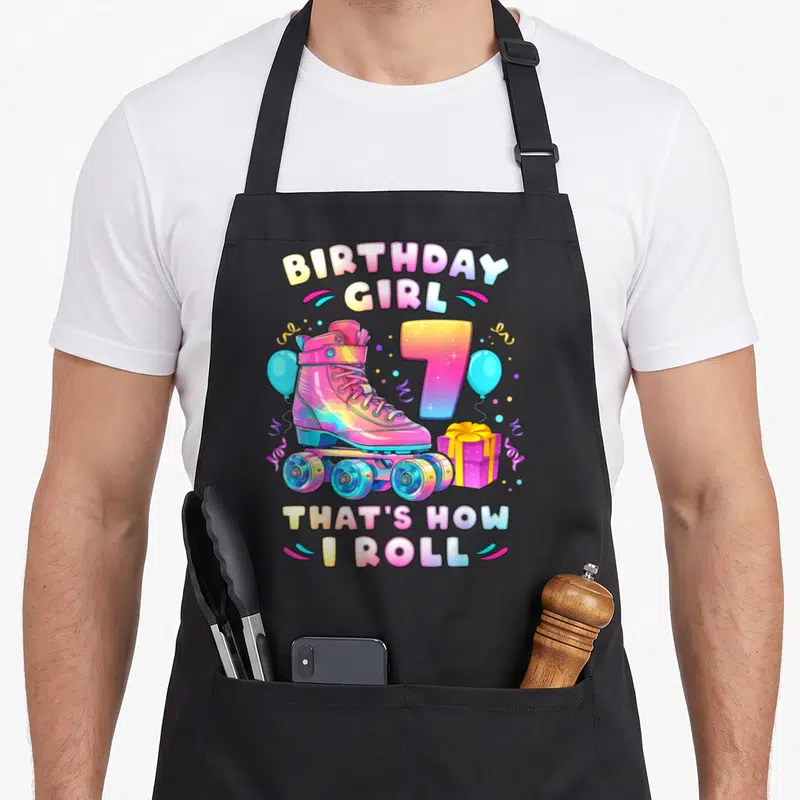 Number birthday apron - Pink roller skate, rainbow number 7, blue balloons, purple gift box, birthday pun text — shown on a black cotton canvas apron against a white background