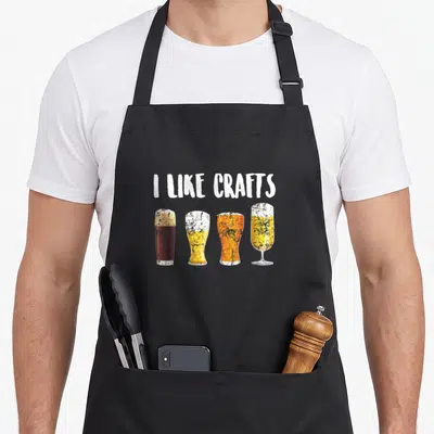 Maker craft apron - I LIKE CRAFTS text above four beer glasses in white yellow orange brown retro style — shown on a black cotton canvas apron against a white background