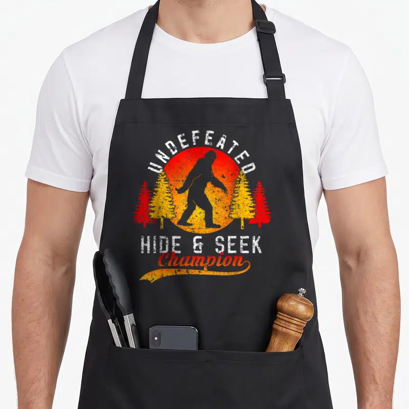 Cute hide bigfoot apron - Grey Bigfoot silhouette against red yellow sun with pine trees and white arched text — shown on a black cotton canvas apron against a white background