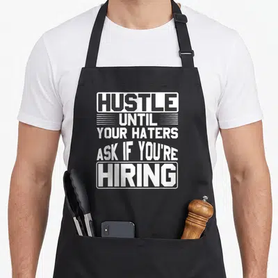 Hater hustle apron - White stacked text reading HUSTLE UNTIL YOUR HATERS ASK IF YOU'RE HIRING with graphic borders — shown on a black cotton canvas apron against a white background